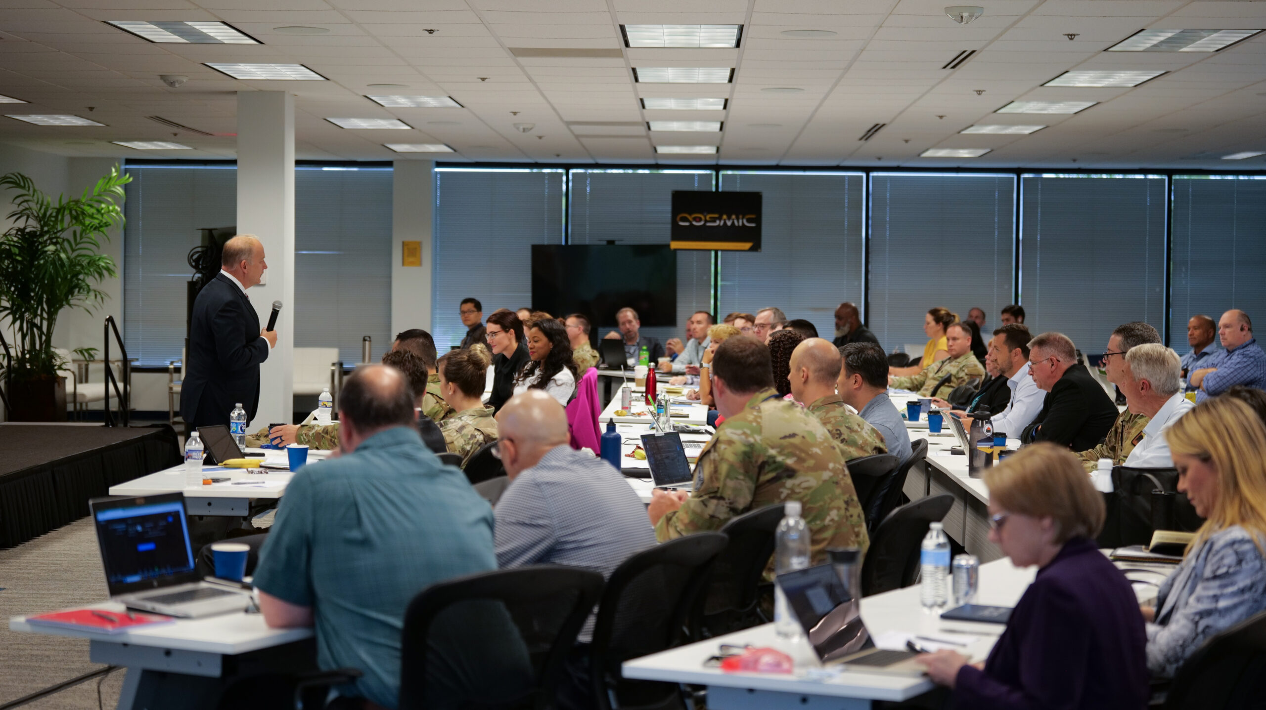 Photo of a speaker delivering a presentation to an audiance of mixed civilian, industry, academia, and DoD attendees in a large conference room.